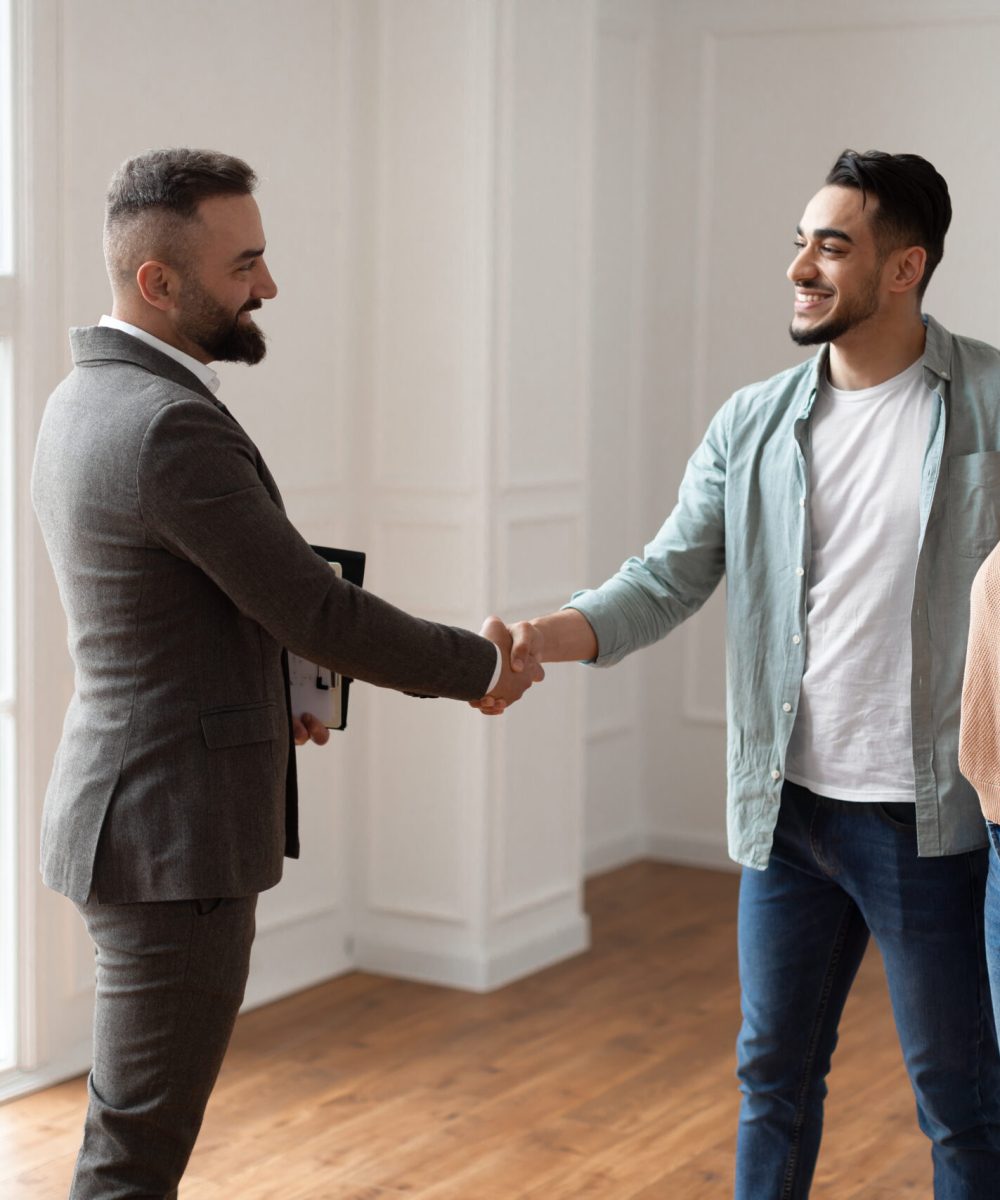 Successful Deal Concept. Portrait Of Young Family Buying New Apartment, Husband Embracing Wife Shaking Hands With Smiling Property Agent. Advisor In Suit Selling Flat To Happy Millennial Buyers