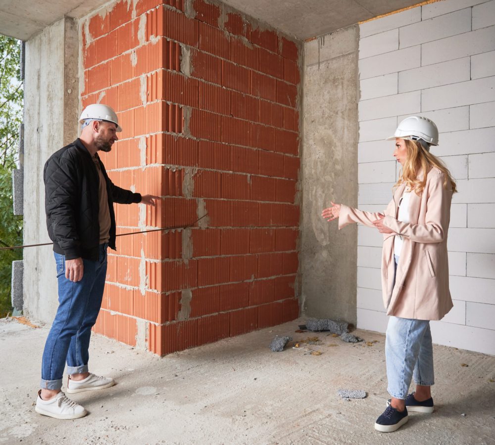 Full length of woman standing by brick wall and discussing apartment replanning and renovation with specialist. Female homeowner having meeting with builder in building under construction.